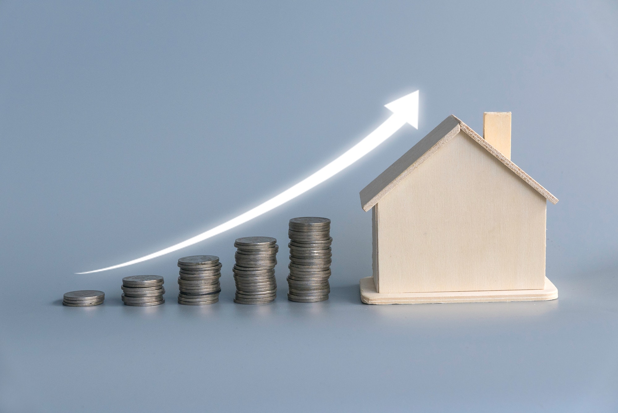 A stack of ascending coins with house model and glowing white arrow on grey background. A stack of ascending coins with house model and glowing white arrow on grey background.